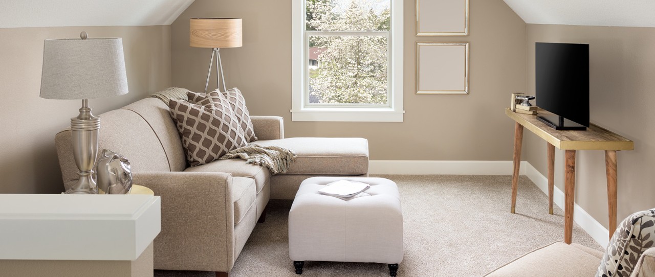 Brightly lit living room with natural light featuring a beige sofa with chaise, white ottoman, and television